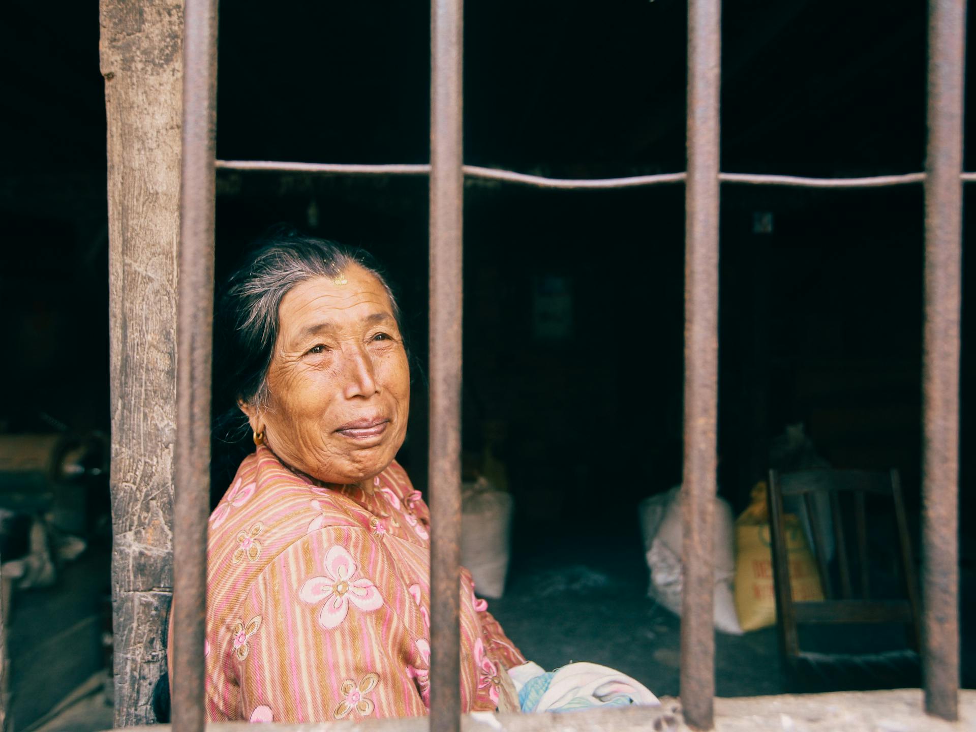 A senior woman in floral attire looking outside through old window bars, bright daylight.