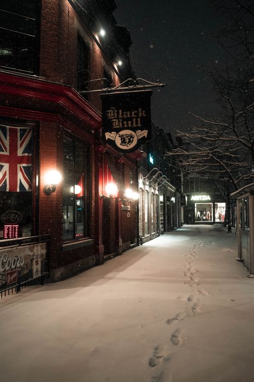 Free A snowy evening street scene of the Black Bull Tavern in Toronto, featuring illuminated signs and footprints in the snow. Stock Photo