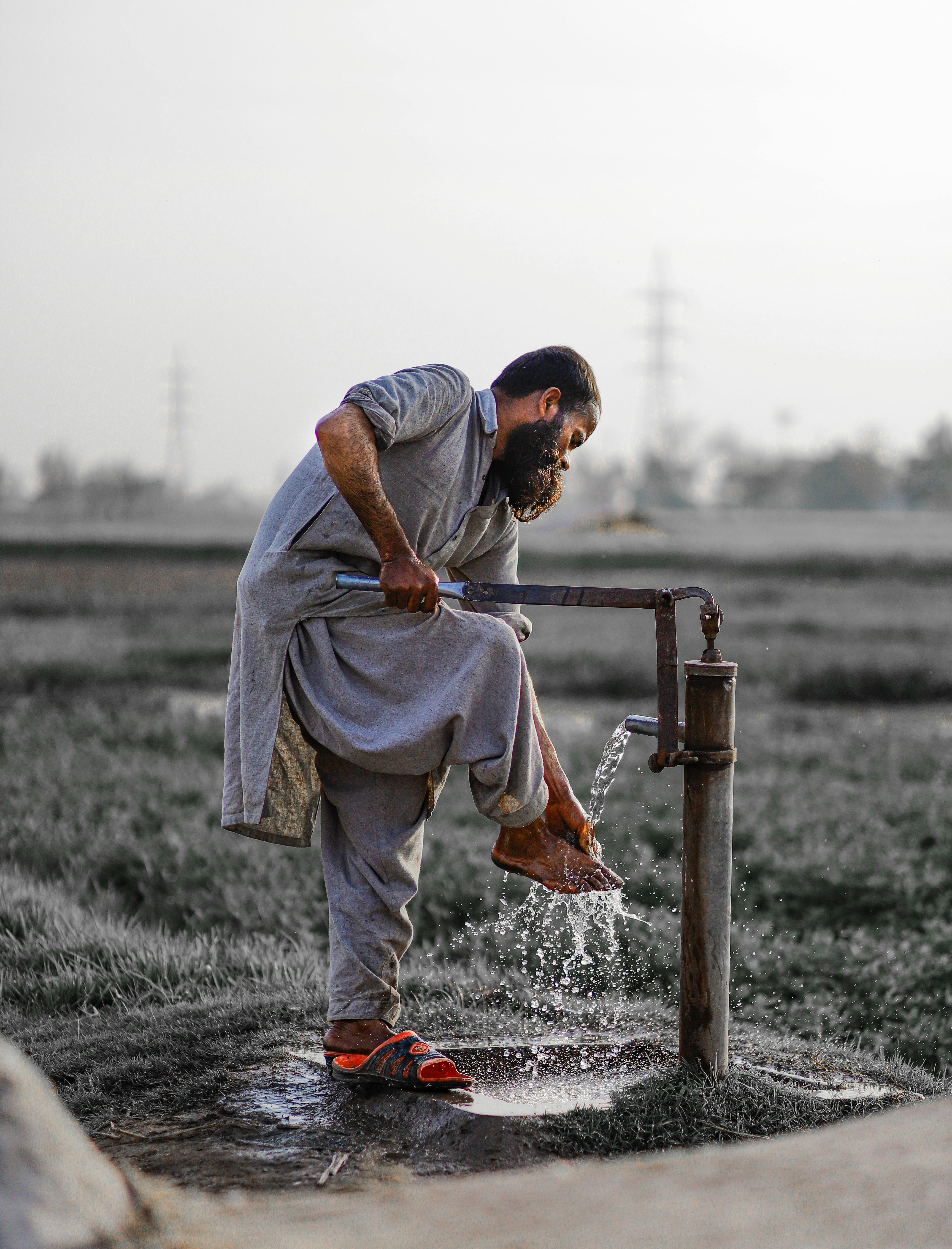 bearded adult man washing his foot with water from pump