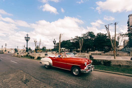 Vintage red convertible driving through a sunny, scenic urban area with cloudy skies.
