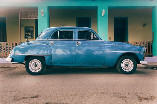 Free A vintage blue sedan parked in front of a vibrant building, showcasing timeless style and color contrast. Stock Photo