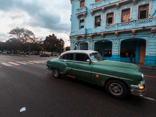 Free Classic green car driving past a colonial blue building on a city street in twilight. Stock Photo