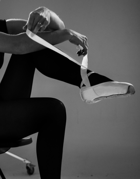 A ballerina sitting and adjusting her pointe shoes in a black and white studio setting.