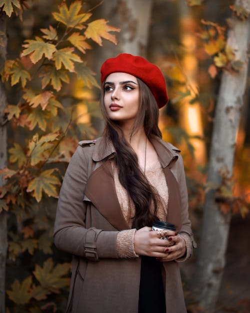 A Woman in Brown Coat and Red Barrette Hat Holding a Cup