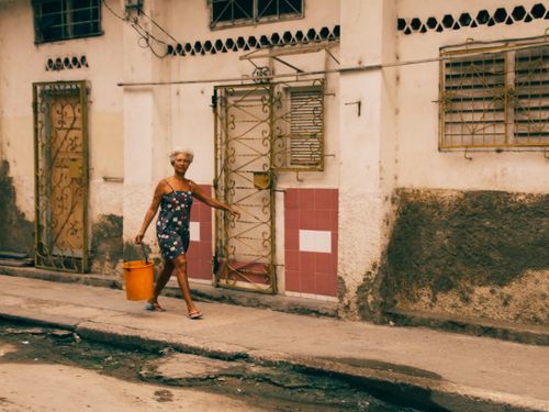 Free Elderly woman carrying an orange bucket, walking past an old urban building facade. Stock Photo