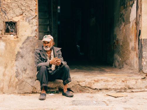 Free An elderly man sits pensively outside a weathered building, capturing a moment of reflection and urban life. Stock Photo