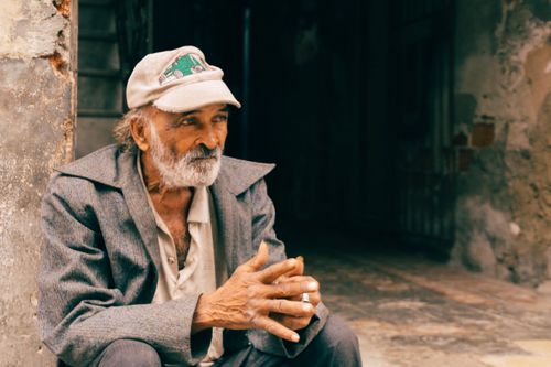 Free Elderly man in a cap sitting outdoors against a rustic wall, thoughtfully gazing. Stock Photo