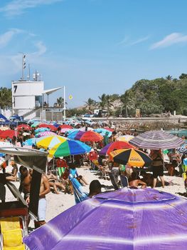 Vibrant crowded beach scene with colorful umbrellas and clear blue sky.