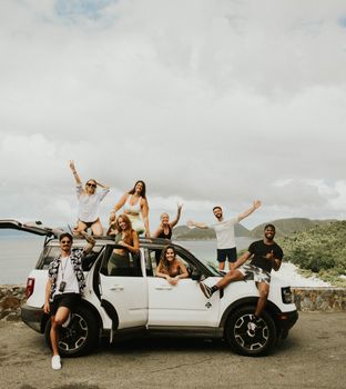 A cheerful group of friends pose together with a car by the sea, celebrating friendship and adventure outdoors.