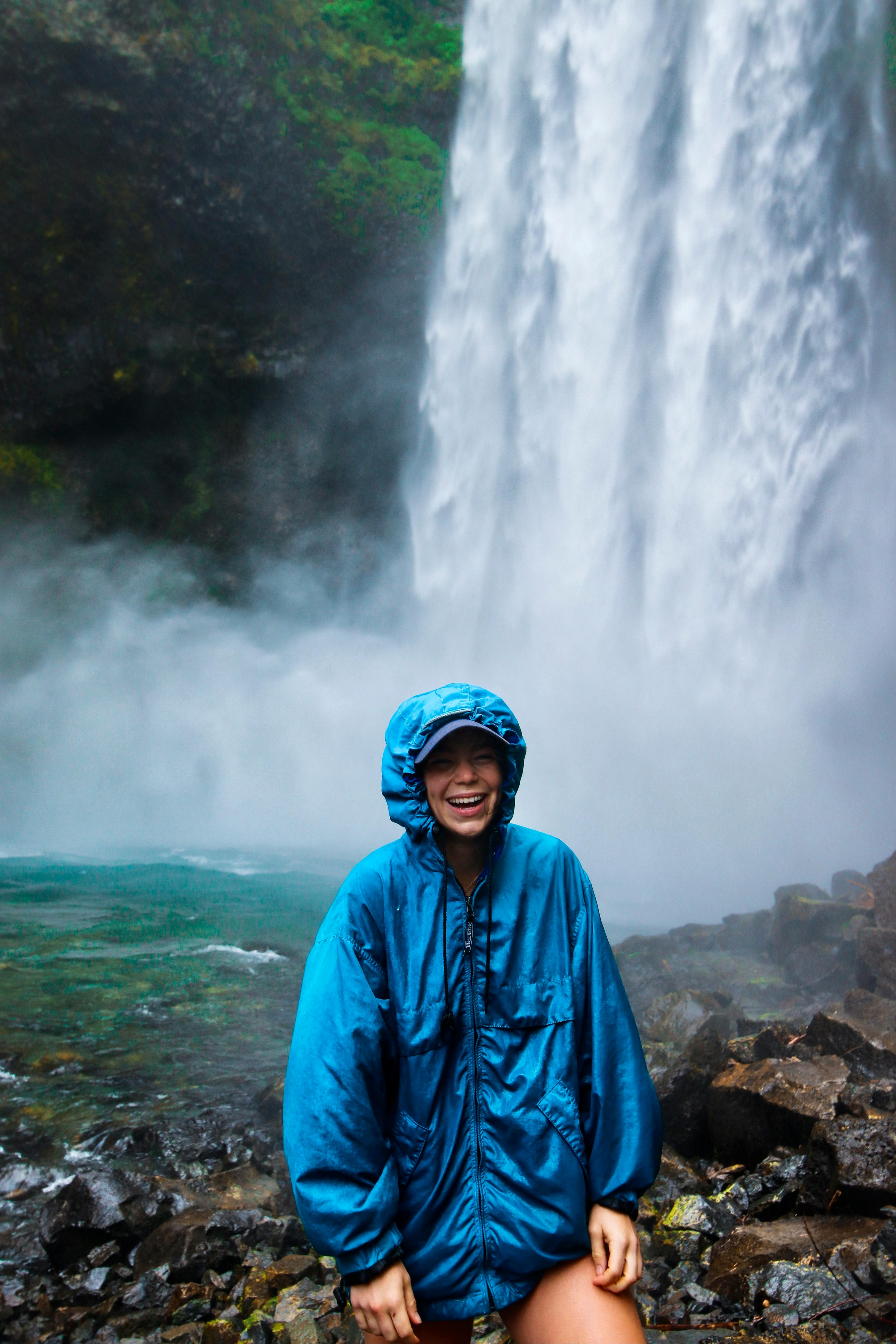 Happy teenager in a blue jacket stands by a stunning waterfall in summer.