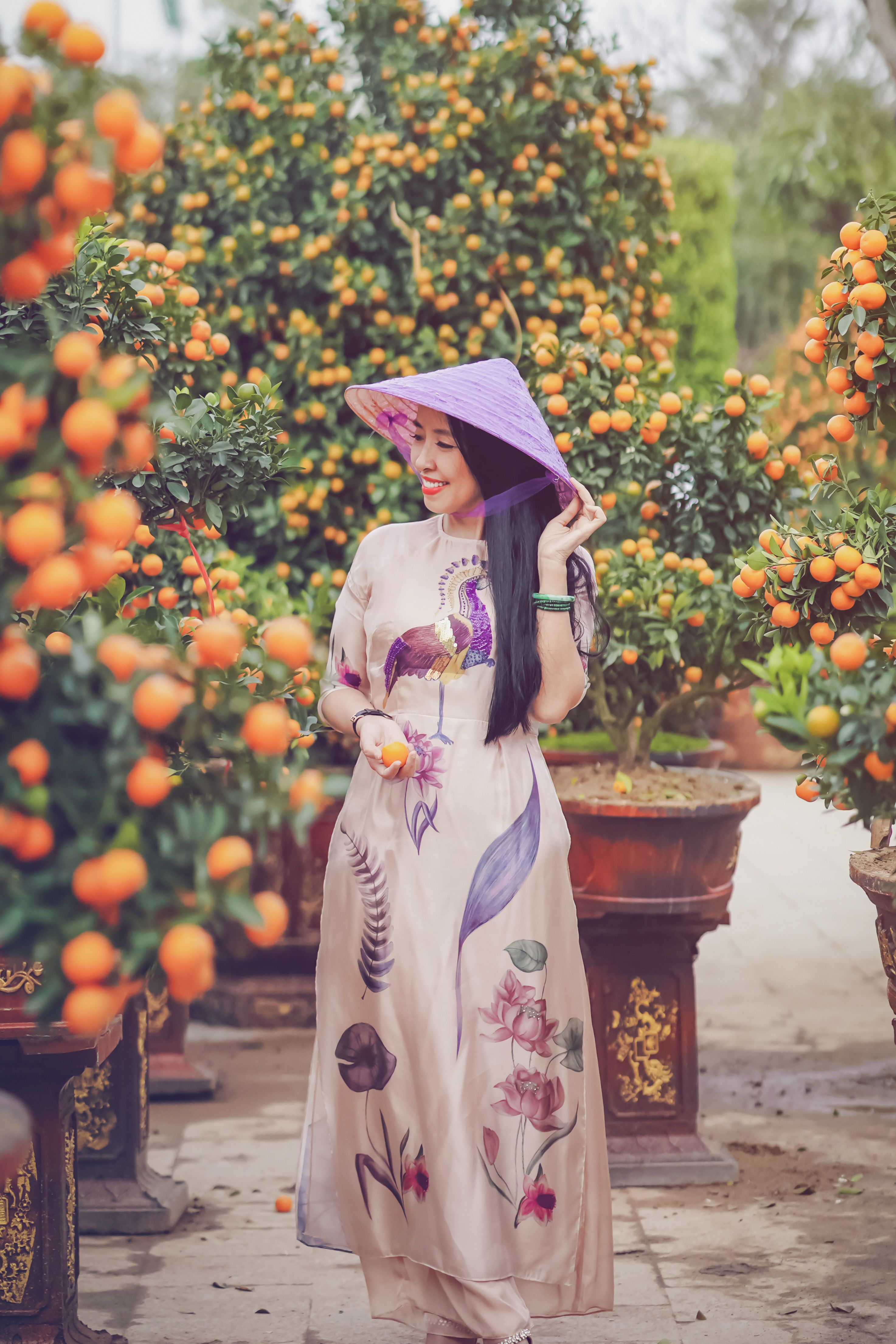 Smiling Asian woman in a floral dress and hat standing among vibrant orange trees.