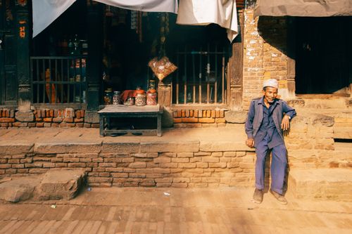 Free An elderly man sits in the sun outside an old brick shop, capturing a moment of daily life. Stock Photo