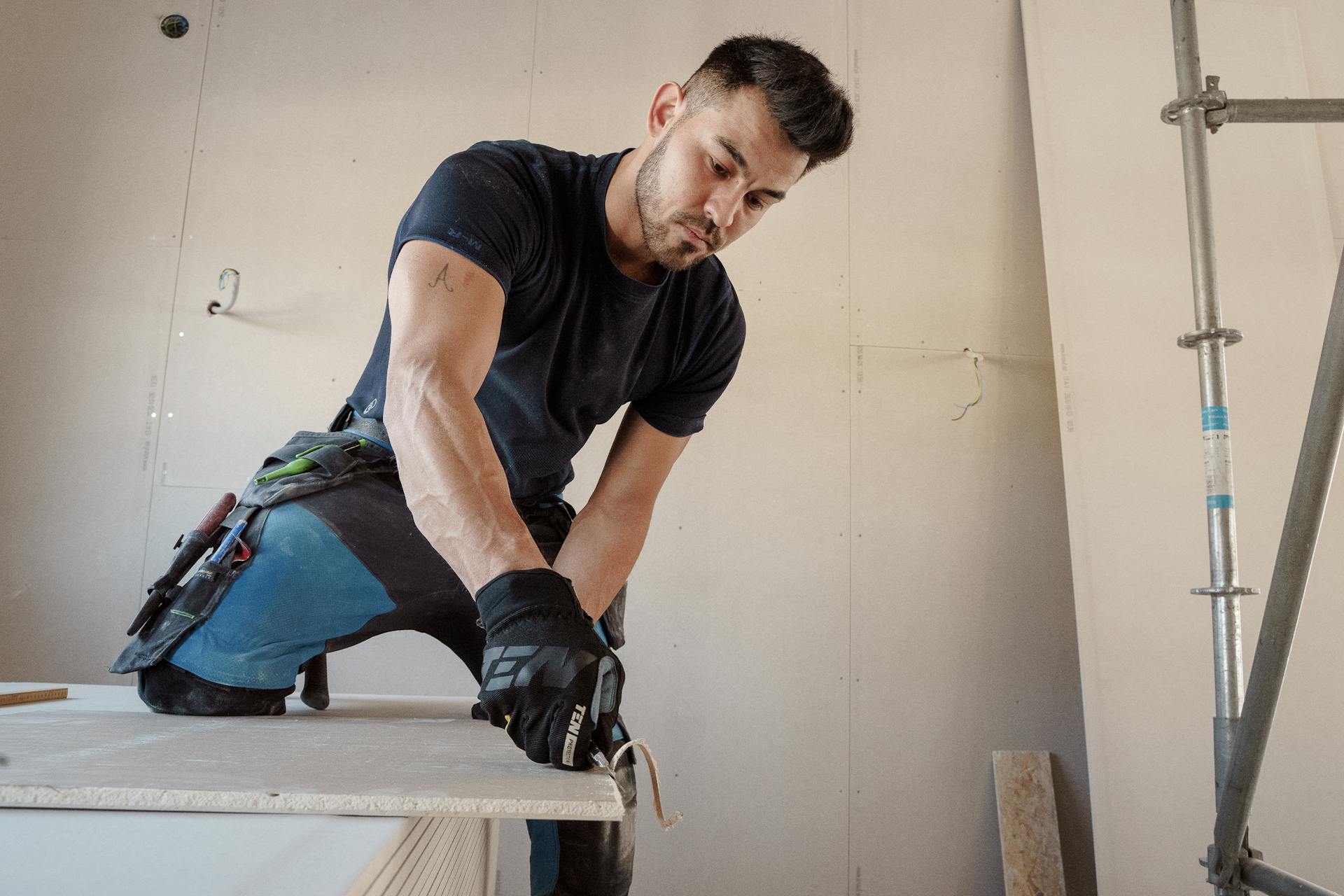 Man in gloves cutting plasterboard for drywall installation inside a building.