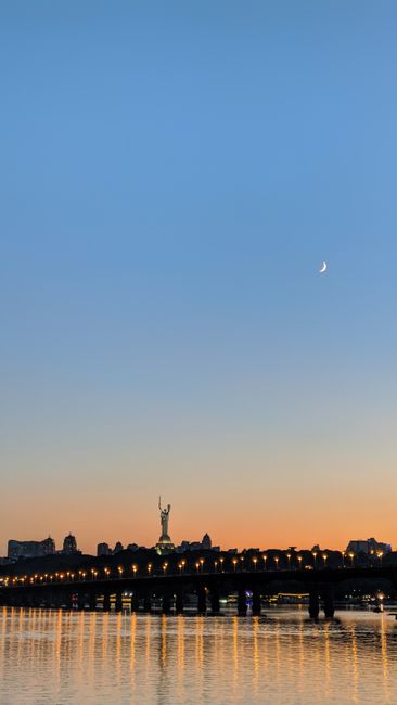 A picturesque view of Kyiv's skyline featuring a bridge and crescent moon against a sunset sky.