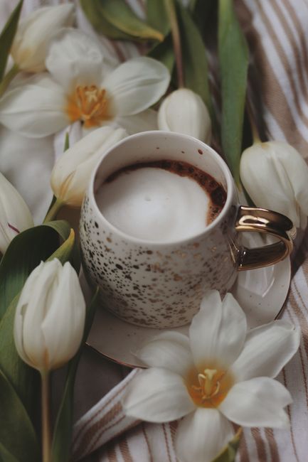Speckled cappuccino cup surrounded by white tulips on striped fabric.