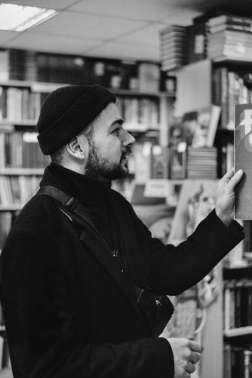 Free Black and white photo of a man wearing a beanie and jacket browsing books in a bookstore. Stock Photo