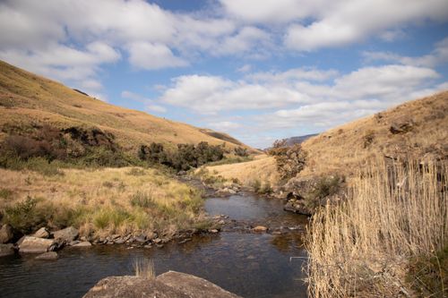 Free Tranquil river flowing through grassy hills with a cloudy sky and warm tones. Stock Photo