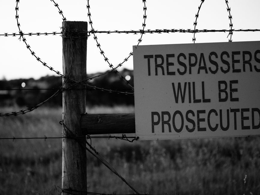 Monochrome image of a barbed wire fence with a sign warning trespassers will be prosecuted.