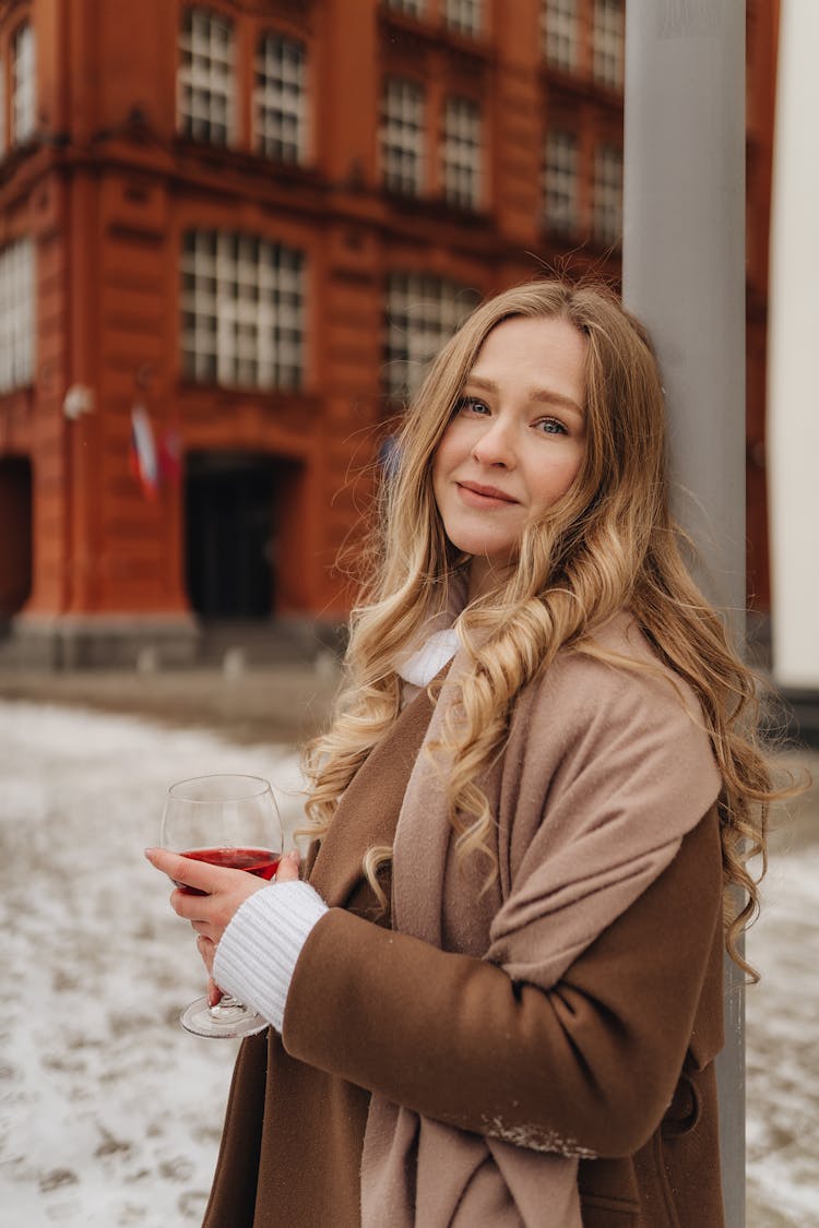 Woman With Glass Of Red Wine In Hand Leaning Against Street Pole