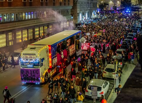 A crowded nighttime protest with banners and a decorated bus on a city street.