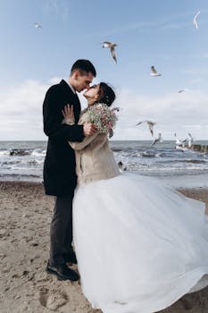Bride and groom share a romantic kiss on a sunny beach as seagulls soar in the background.