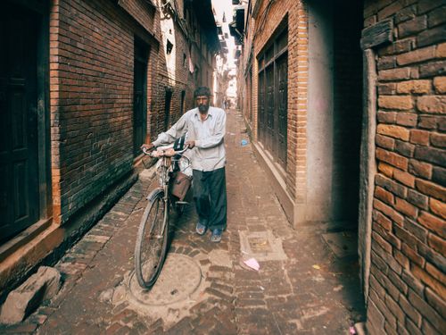 Free An adult man walking his bicycle through a narrow brick alley between tall buildings. Stock Photo