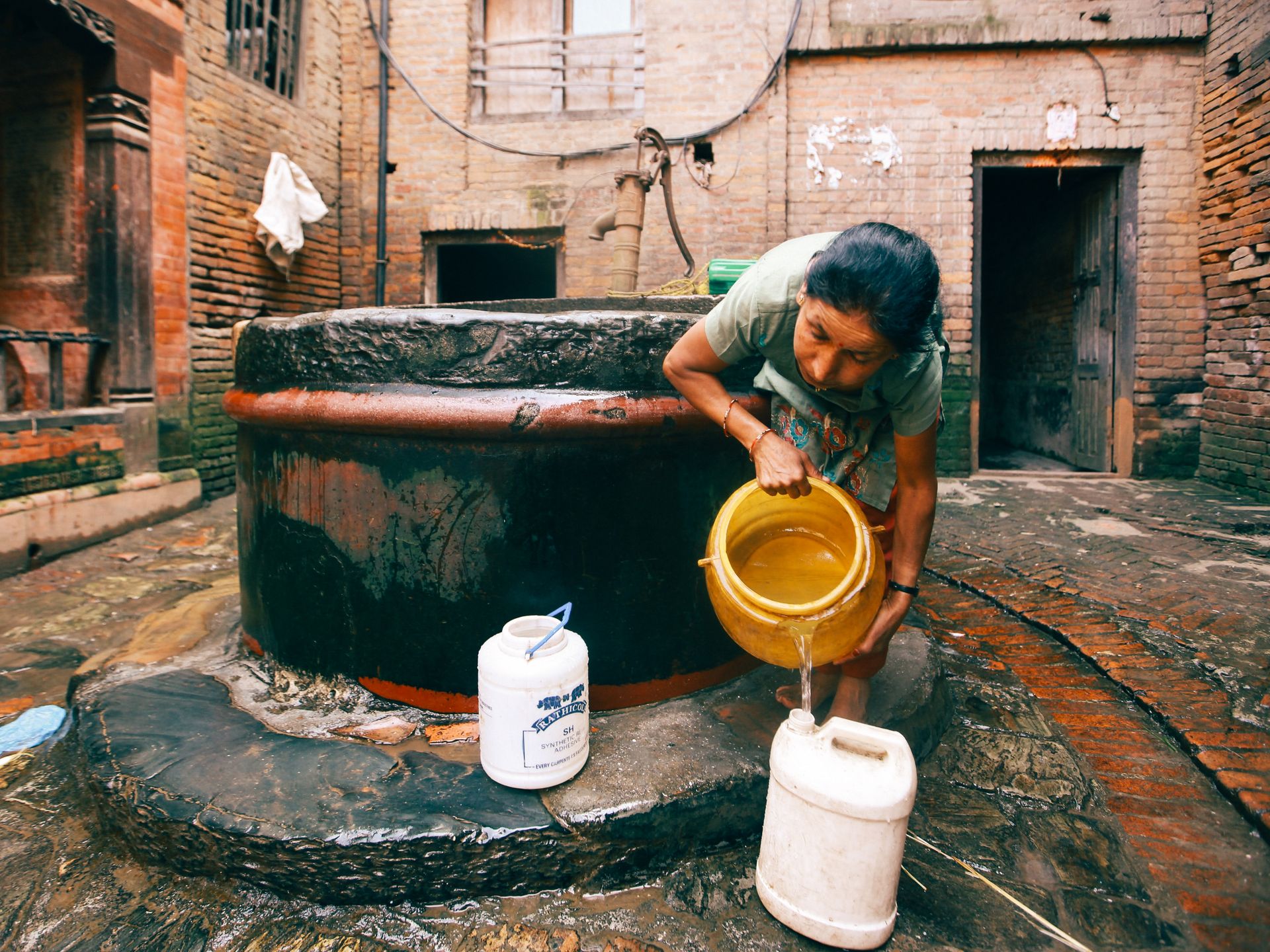 An adult woman gathers water from an outdoor well in a rustic courtyard.