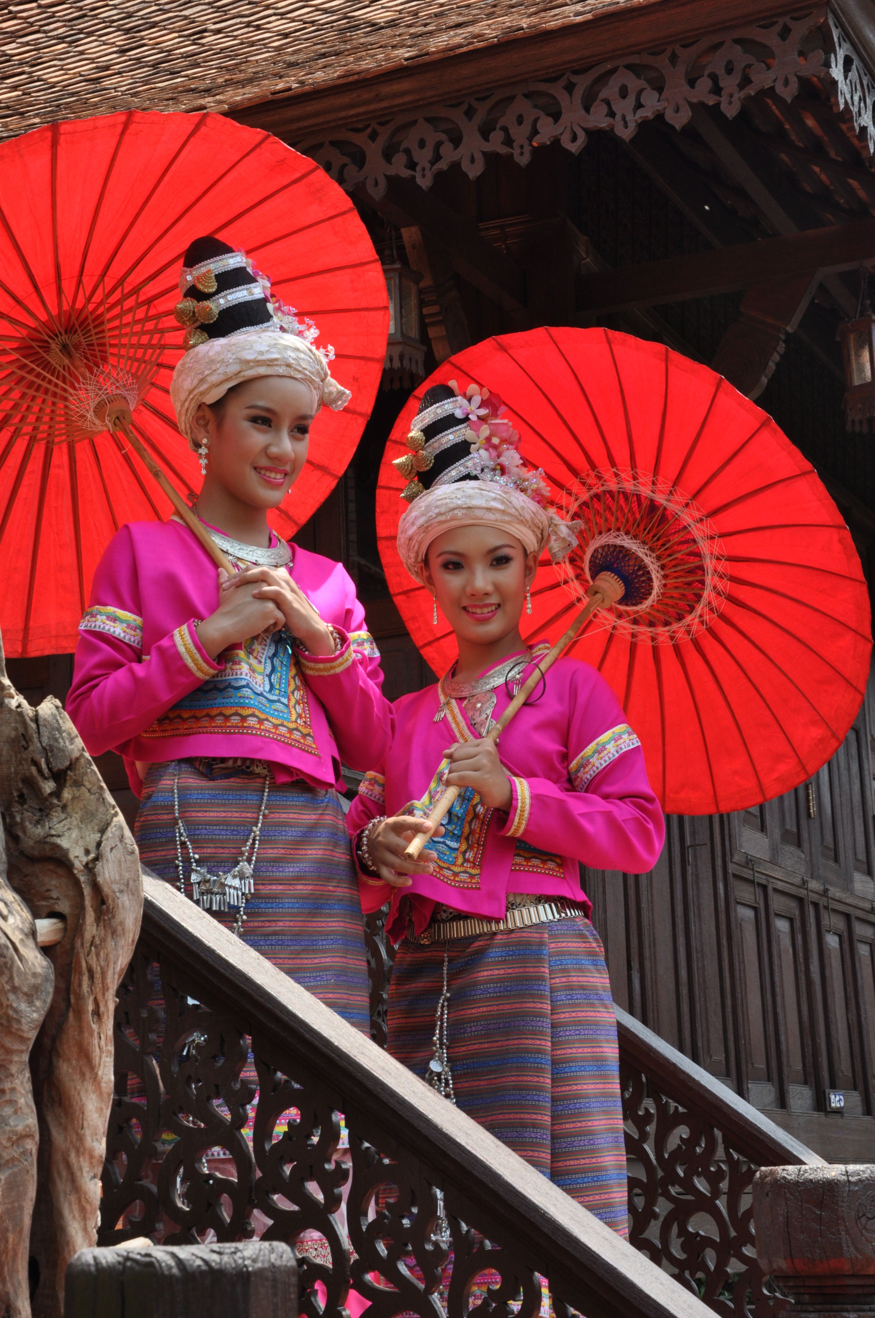 Two young women in traditional Thai clothing with red umbrellas posing outdoors.