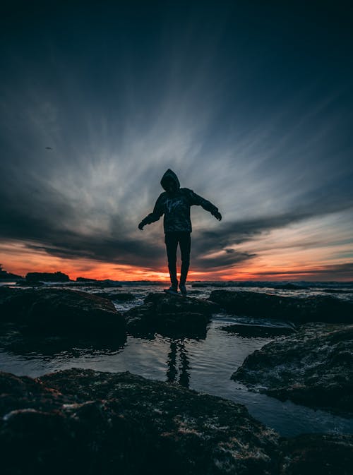 Free Person Wearing Hoodie Standing on Rock Surrounded by Body of Water Stock Photo