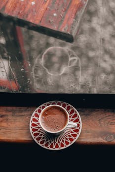 A warm cup of coffee on a windowsill with rain-drenched glass in the background, evoking coziness.