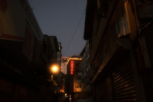 Atmospheric city street at dusk with neon hotel sign and warm lights casting shadows.