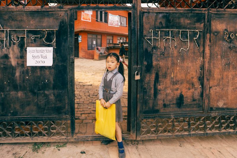 A schoolgirl in uniform holds a yellow bag at a school entrance in Nepal.