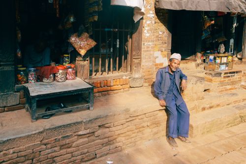 Free Charming image of an elderly man resting outside a rustic brick shop in a sunny town. Stock Photo