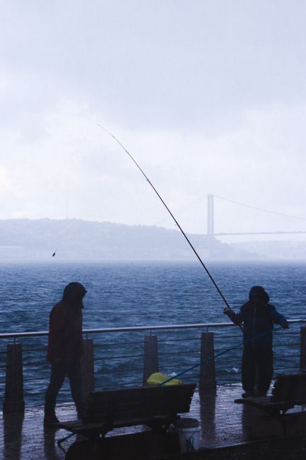 Two adults fish on a foggy day by the Bosphorus Bridge in Istanbul, Turkey.