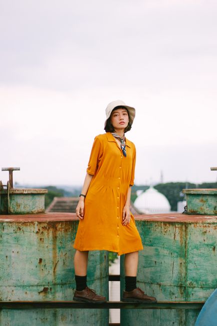 Woman wearing a yellow dress and hat posing outdoors against industrial backdrop.