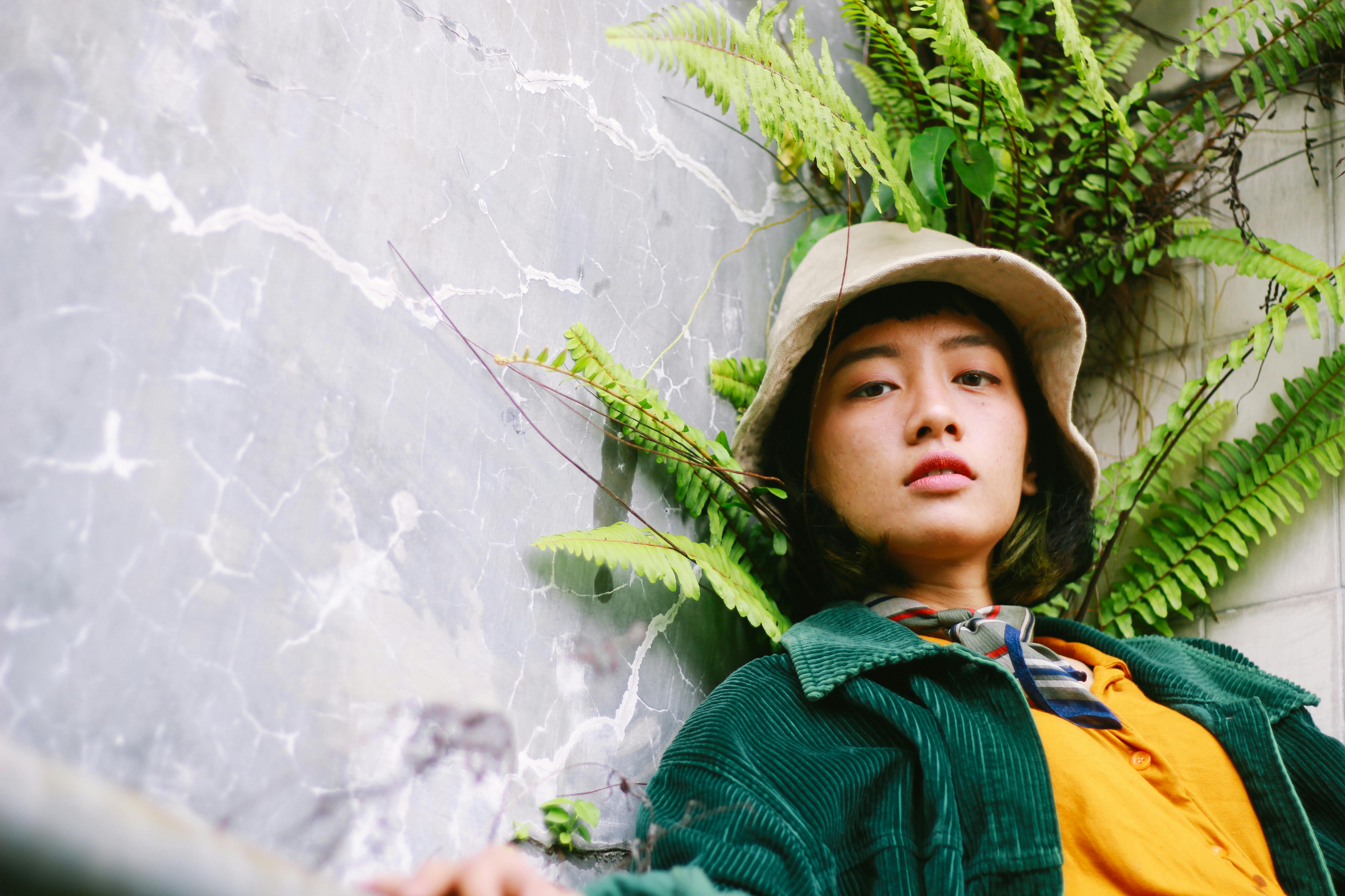 Portrait of a stylish woman in a hat, leaning against a wall with ferns.