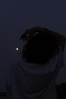 A woman silhouettes against a dark night sky, gazing at the moon.