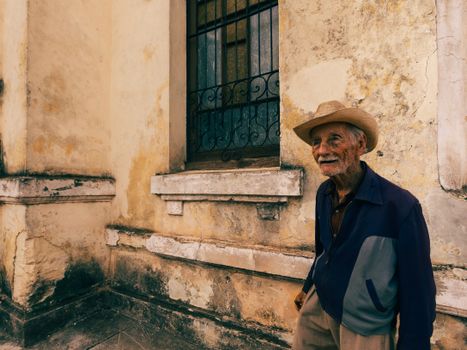 Portrait of a smiling elderly man wearing a hat, standing by a rustic yellow wall with a window.