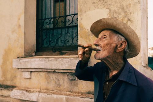 Free An elderly man in a hat enjoying a cigar outside a rustic building, capturing vintage charm. Stock Photo
