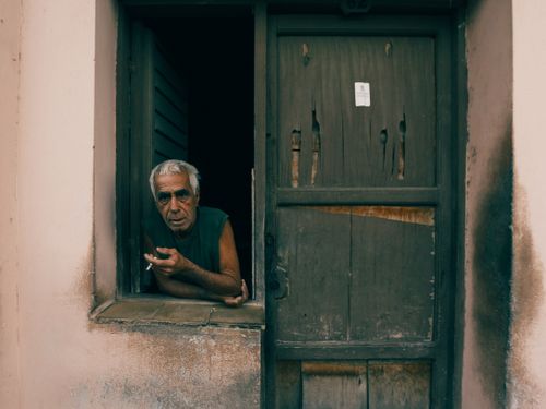 Free Senior man smoking, leaning on a rustic window. Thoughtful portrait in an aged setting. Stock Photo