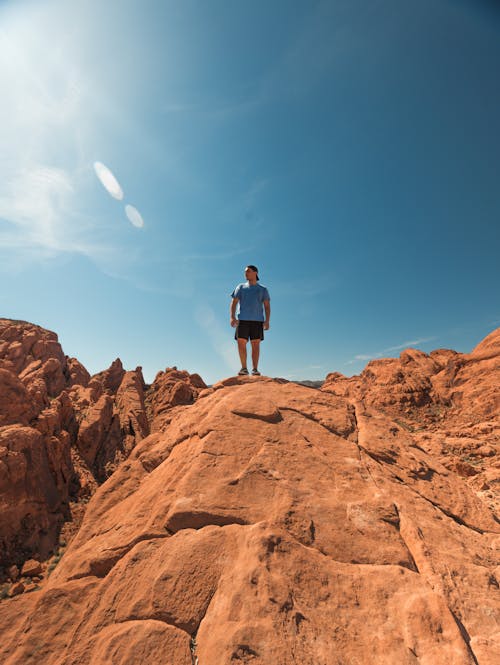 Man Wearing Blue Shirt and Black Shorts Standing on Top of Brown Rock Formations Under Clear Sky