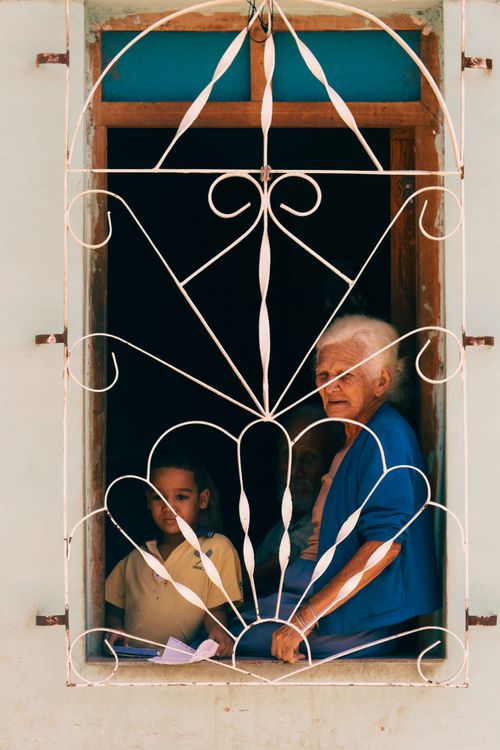 Free A senior woman and a child look out from a window with ornate metalwork, capturing a moment of connection. Stock Photo