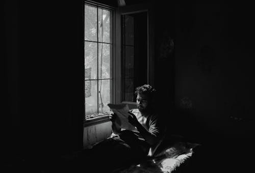 Grayscale Photo of Man Reading By a Window