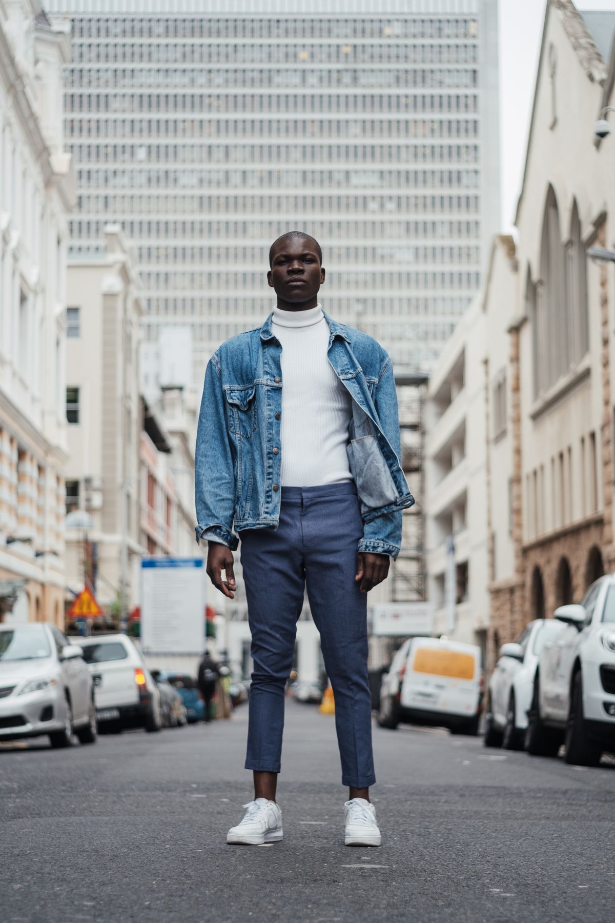 A man in a denim jacket and white turtleneck standing confidently on a city street
