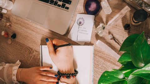 Woman's hand writing in a journal surrounded by crystals, plants, and laptop on a wooden desk.