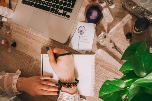 Free Woman's hand writing in a journal surrounded by crystals, plants, and laptop on a wooden desk. Stock Photo