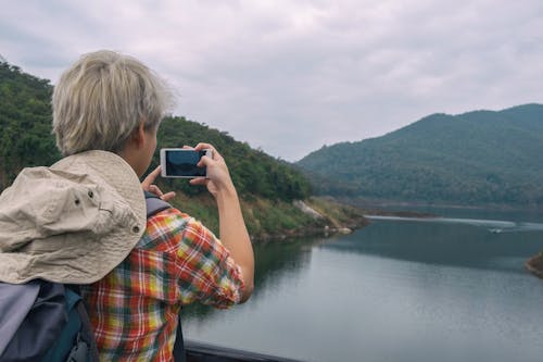 Person Holding Smartphone While Taking Photo of Calm Water Near Mountain at Daytime