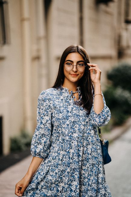 Portrait of a smiling woman with glasses, wearing a blue floral dress on a city street.