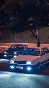 Vintage cars with headlights on parked on a quiet street at night under streetlights.