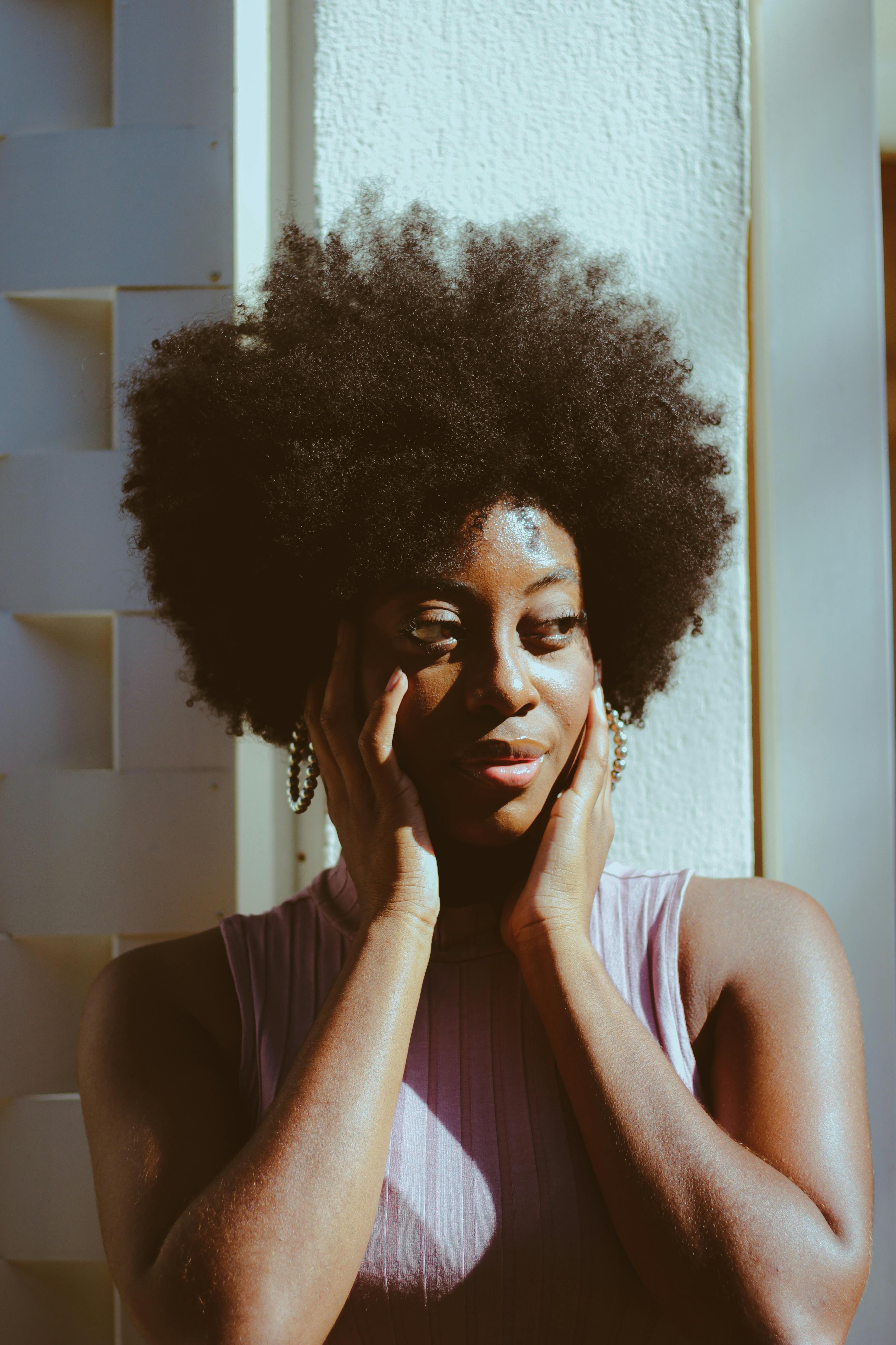 Close-up portrait of a woman with afro hair basking in sunlight, hands on face.